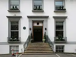 A flight of stone steps leads from an asphalt car park up to the main entrance of a white two-story building. The ground floor has two sash windows, the first floor has three shorter sash windows. Two more windows are visible at basement level. The decorative stonework around the doors and windows is painted grey.