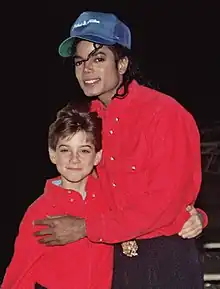 A smiling Jackson wears a blue baseball cap and a red shirt. On his left, a young boy smiles. He is dressed in a red shirt, too.