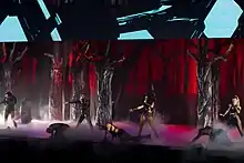 Little Mix performing in black and white outfits on stage with a forest backdrop.