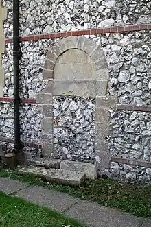 Part of a flint wall with red-brick bands, in which a tall arch-shaped blocked entry is set.  It has flints and stone blocks, and is surrounded by stones of different sizes.