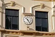 Close-up photograph of a wall-mounted clock with a white face and black Roman numerals, on a pastel-yellow wall.