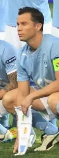 A man with short dark hair kneels on a football pitch  before a match, wearing a light blue shirt, white shorts and light blue socks. He is holding a pennant.