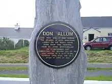 Black circular plaque mounted on a standing rock. Text reads: "Don Allum, the first man to row across the Atlantic in both directions, landed on the rocky shore down from this memorial, from St. John's Newfoundland on 4th September 1987, completing the second leg of his epic voyage in a 19 ft. 10 ins. plywood open boat, the QE 3, after 77 days at sea."