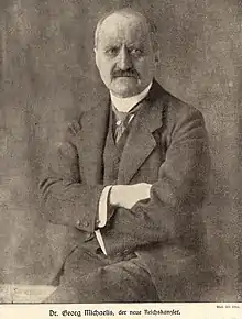 Black and white photo of an elderly man sitting in a suit with folded hands, mustache and bald head.