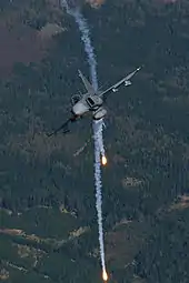 Head on view of fighter jet banking right while releasing flares against a background of green woodland