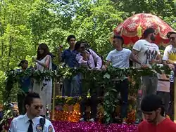 A parade float with fencing is decorated with flowers. Multiple men and women of South Asian descent lean towards fans on the street.