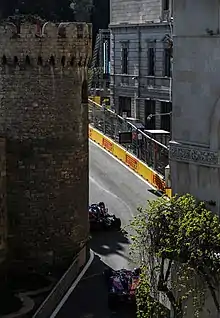 A tarmac race course, lined with concrete and metal barriers, passes between a building and a turret.