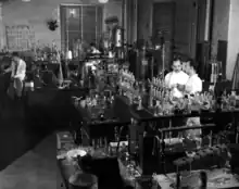 Photograph of a room with multiple scientists in white lab coats standing among tables covered in glassware