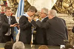 A smiling Macri puts on a blue-and-white sash with the help of Federico Pinedo and two other men
