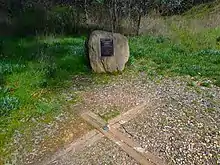 A boulder with a plaque is in the background, with a wooden cross embedded in the ground in the foreground.