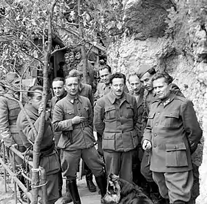 a black and white photograph showing half a dozen men standing on a narrow wooden walkway alongside a rock wall