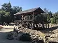 Historic photograph of Sutter's Mill in Coloma in 1850. It is a largely open-walled wooden building set on stits among sparsely vegetated mountains.