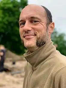 A partially-bald white man with mutton chops wearing a beige fleece smiling at the camera.