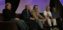 Five older men sit on a couch, with an arched, decorative purple background behind them. Ralston, leaning back in his seat, has short hair and wears a shirt with slacks.