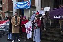 Several protesters holding banners, Gosek-Popiołek speaking to a microphone, "Left Together" flag visible behind her