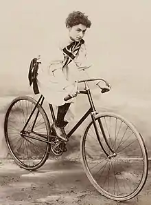 White woman posed on a bicycle, wearing a baggy white garment with a Union Jack on the chest