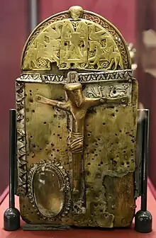 A hand-bell shaped shrine with a main face containing large central crucifixion, and a rock crystal to the lower left. The embossed panel above Christ's right arm is decorated with animals. The semicircular cap and crest above the main face shows a highly detailed central figure, likely an ecclesiastic, holding a book. Horse riders and birds are on either side of him.