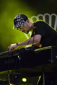 Shuya Okino of Kyoto Jazz Massive bending over a keyboard onstage, wearing a t-shirt, sunglasses, and ball cap
