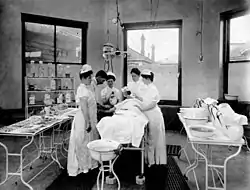 In the centre is a bed on which is a patient covered in a sheet. To the right of the patient are two female nurses, and to the left are two female nurses flanking a male doctor, all facing the patient. Flanking the patient and surgical team are four tables, two on each side. The tables on the left contain an array of metal surgical tools, the tables on the right various pans and covered pitchers.Behind can be seen three of the eight walls of the octagonal room, each centred by a large window. through the central window can be seen the roof and walls of another building, as well as several chimneys. In front of the leftmost window is a metal stnad with three shelves containing various large glass bottles.