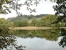A view across Loch Arthur shows a body of water in the foreground reflecting trees on the other bank. A hill rises in the distance.