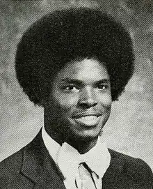 A Black teen is wearing a dinner jacket and white bow tie; with a large afro, he is facing right of the camera and smiling.