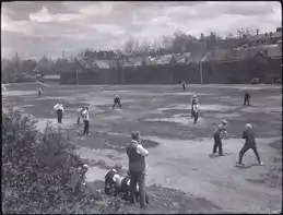 Men playing baseball. There are buildings, automobiles, and hydro lines in the background.